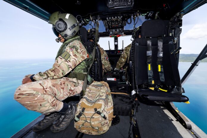 Two Army soldiers in camouflage uniforms sit inside a military helicopter cockpit, with one facing the open side and a large backpack nearby, flying over water and coastline.