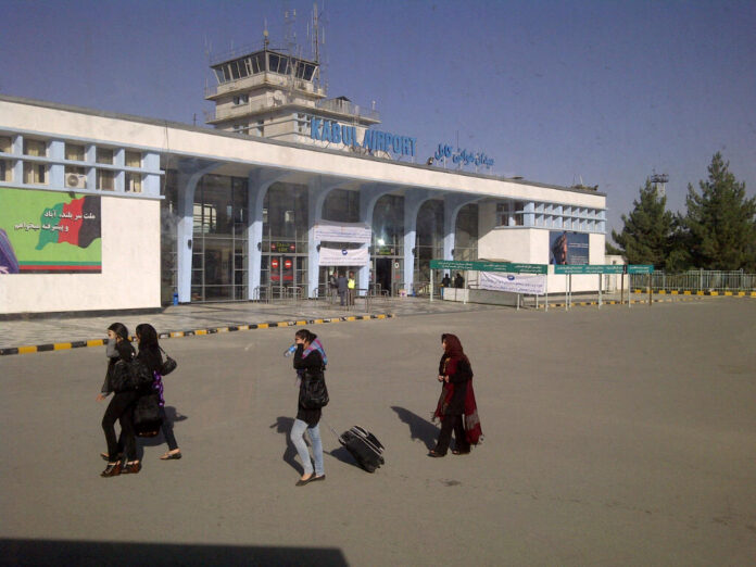 People walk outside the main entrance of Kabul Airport on a sunny day, with a control tower visible on the roof and signage in English and Dari, as Army pathfinders coordinate security nearby.