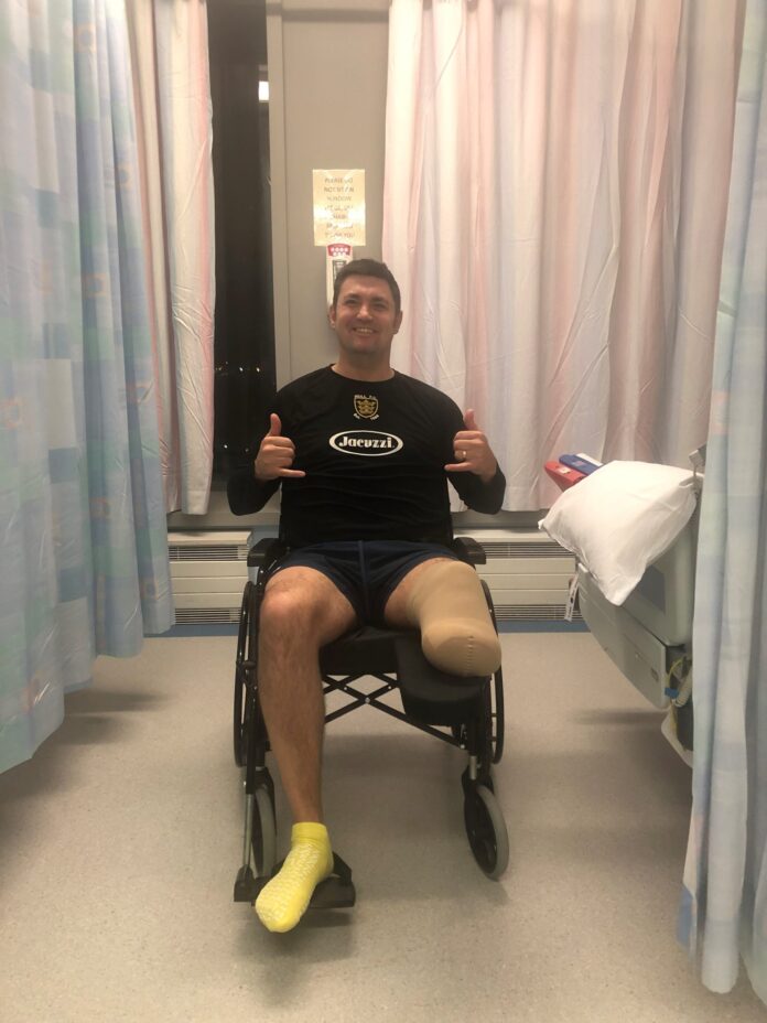 A man sits in a wheelchair in a hospital room, giving two thumbs up. Wearing a black shirt and yellow socks, this military pathfinder remains positive after above-the-knee amputation.