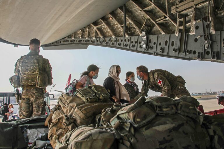 Army soldiers assist civilians boarding a military aircraft, with gear bags in the foreground and the plane’s ramp open on an airfield.