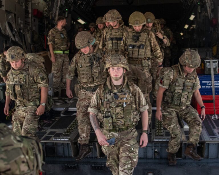 A group of Army soldiers in camouflage uniforms and helmets walk out from the rear of a military aircraft.