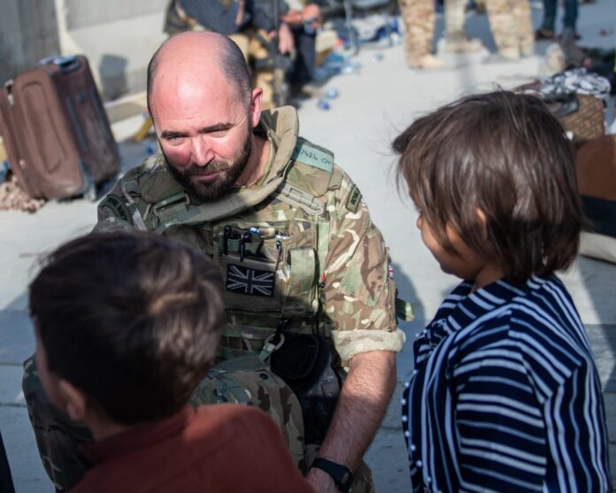 A veteran in British military uniform kneels and interacts with two children outdoors, with luggage and other people—perhaps amid resettlement—visible in the background.