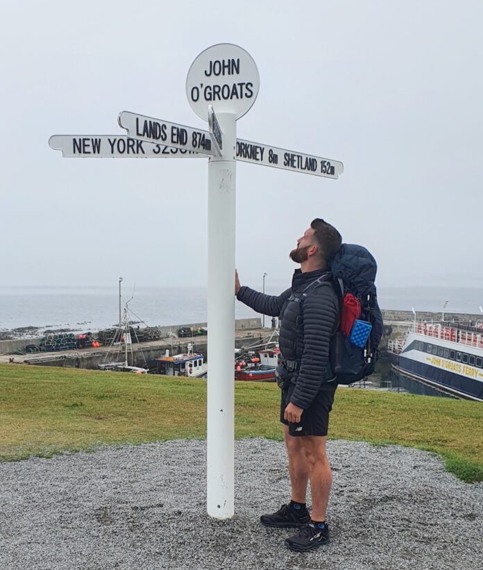 A man with a backpack, perhaps a pathfinder on a resettlement journey, stands next to the John O'Groats signpost, gazing up at the signs under a cloudy sky with the harbor in the background.