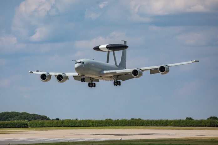 A military surveillance aircraft with a radar dome on top is landing on a runway, showcasing advanced Army technology against a backdrop of trees and blue sky.