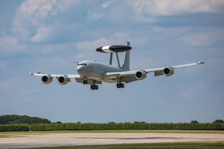 A military surveillance aircraft with a radar dome on top is landing on a runway, showcasing advanced Army technology against a backdrop of trees and blue sky.