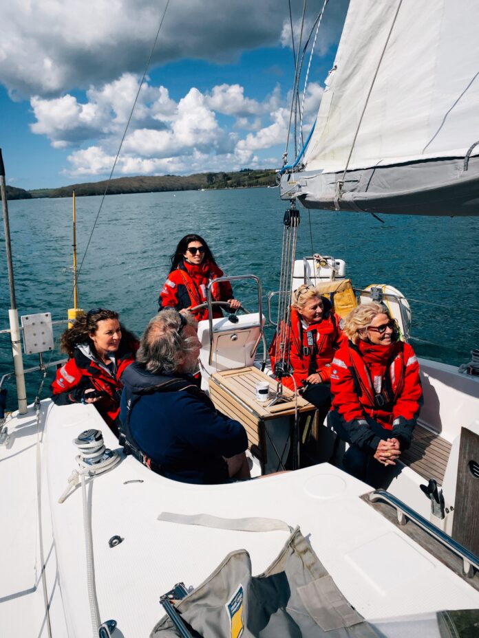 Five people in red jackets, part of an Army resettlement program, sit and stand on a sailboat facing forward, with water and shoreline visible behind them under a partly cloudy sky.