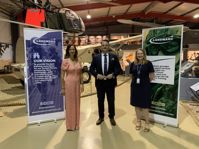 Three people stand indoors between two Landmarc banners, in front of a helicopter display at a museum—highlighting support for Royal Navy veteran resettlement.