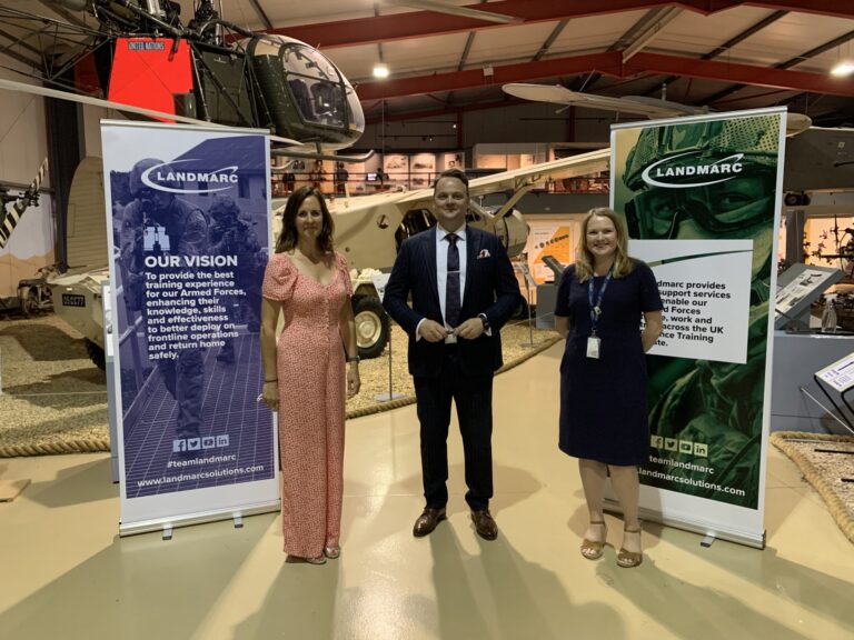 Three people stand indoors between two Landmarc banners, in front of a helicopter display at a museum—highlighting support for Royal Navy veteran resettlement.