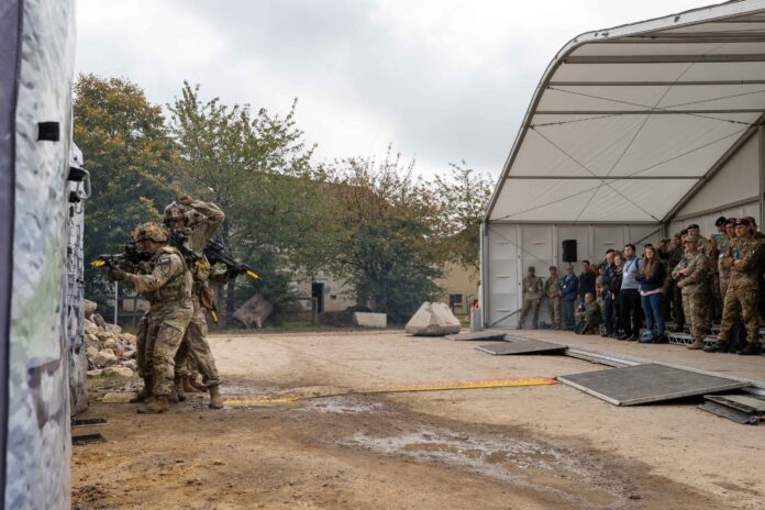 Two soldiers in uniform conduct a tactical demonstration outside a tent, while a group of soldiers, veterans, and civilians observe from inside.