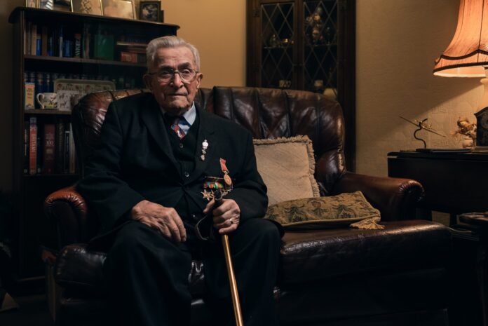 An elderly Army veteran in a suit with medals sits on a leather armchair in a warmly lit living room, holding a cane.