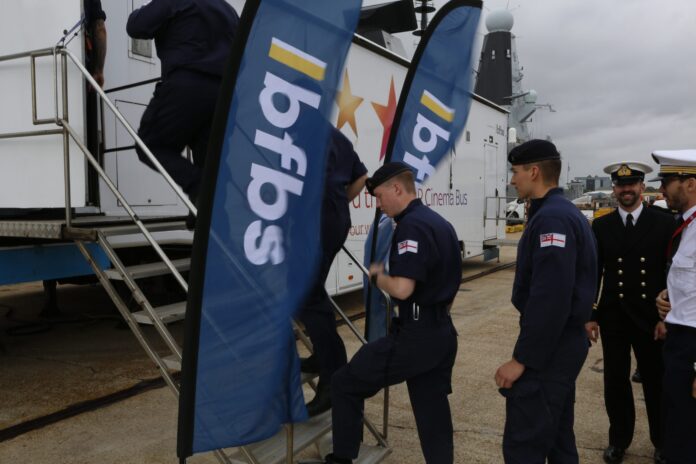 Several uniformed naval personnel, including a Pathfinder veteran, ascend a metal staircase next to two blue BFBS banners, with other officers standing nearby on a cloudy day.