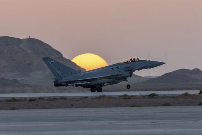 A military jet, part of the Army Pathfinder unit, takes off from a runway at sunset, with mountains in the background and the sun partially hidden behind a hill.