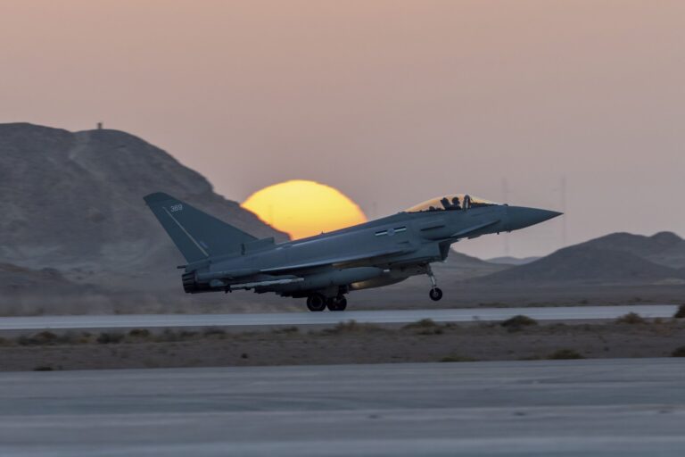 A military jet, part of the Army Pathfinder unit, takes off from a runway at sunset, with mountains in the background and the sun partially hidden behind a hill.