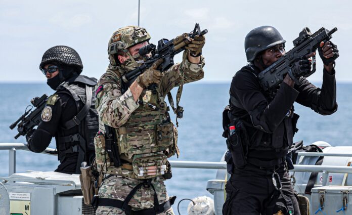 Three armed soldiers in tactical gear, part of the Royal Navy, aim their weapons while standing on a vessel at sea, focusing intently on something off-frame.