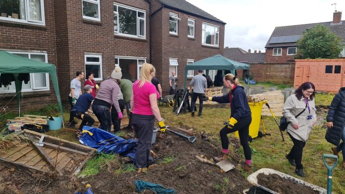 A group of people, including members of the Army, work together in a garden area outside brick buildings, using tools and clearing debris, with canopies and equipment set up nearby.