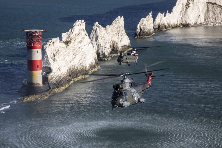 Two Royal Navy military helicopters fly over the sea near a red and white lighthouse and jagged white cliffs, their rotor wash stirring the water's surface.