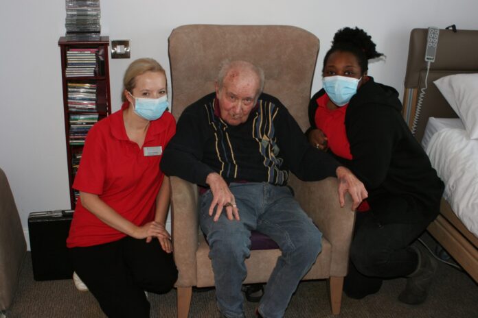 An elderly Royal Navy veteran sits in a chair between two caregivers wearing red shirts and face masks, posing for a photo in a care facility room.