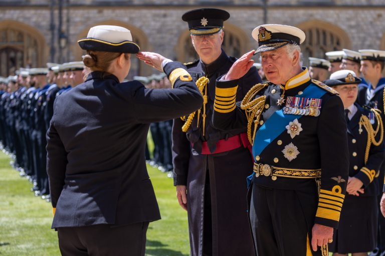 A uniformed officer salutes a decorated Royal Naval officer outdoors, with rows of uniformed personnel standing in formation in the background, marking a moment of Honours.