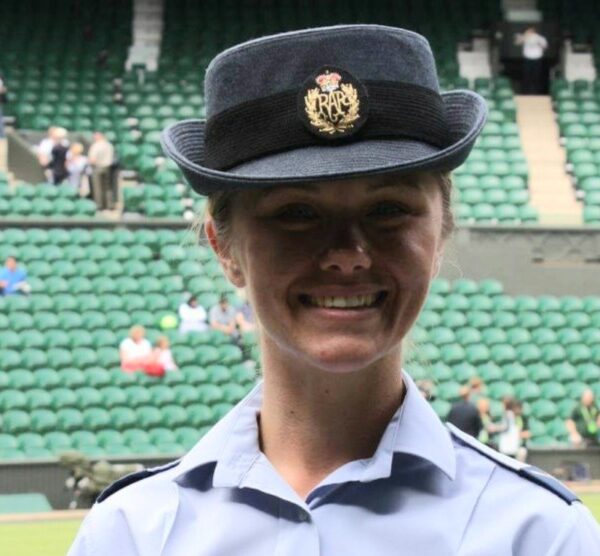 A person in a uniform with a hat featuring a badge is smiling, standing in a stadium with green seats, embodying the spirit of helping through their support for the Royal British Legion.