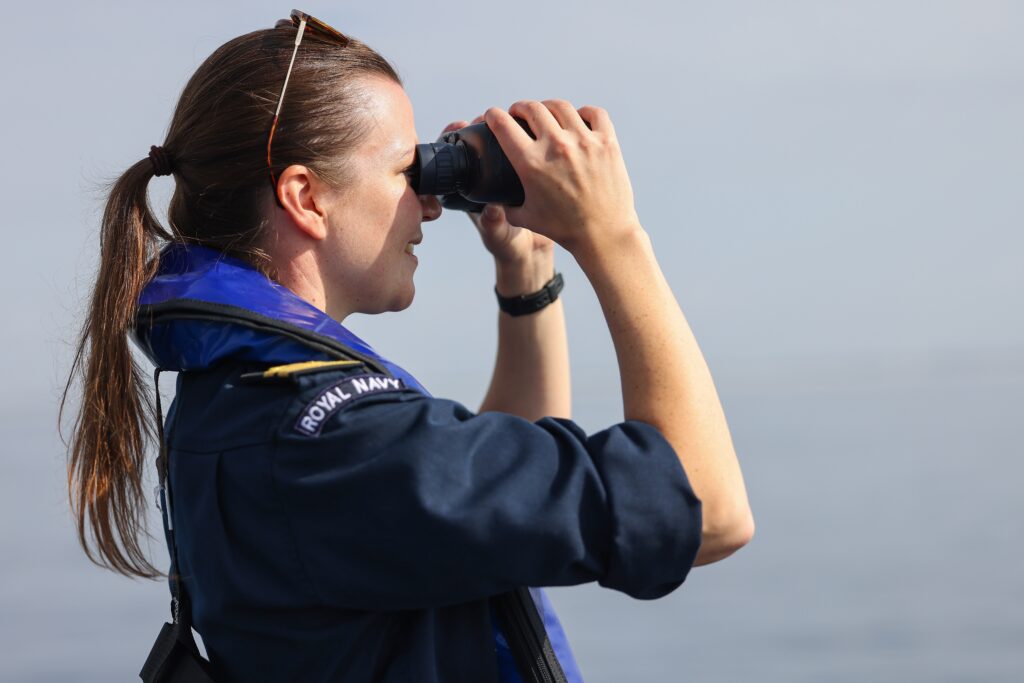 A person in a Royal Navy uniform peers through binoculars, scanning the vast ocean for uncrewed vessels 10,000 miles away.