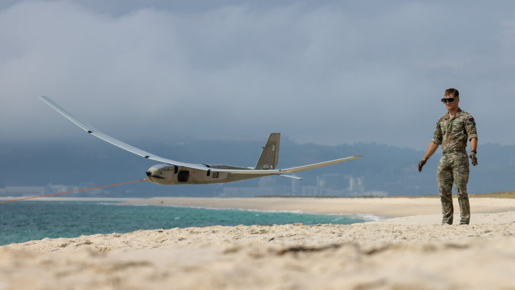 Under a cloudy sky on a sandy beach, a soldier from the Royal Navy operates an uncrewed drone vessel.