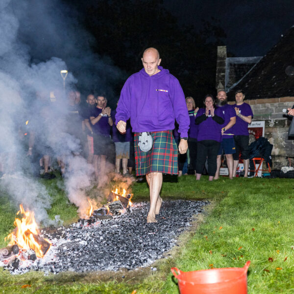 A person walks barefoot on burning coals, feeling the heat with every step, while a group of people watches under the night sky.