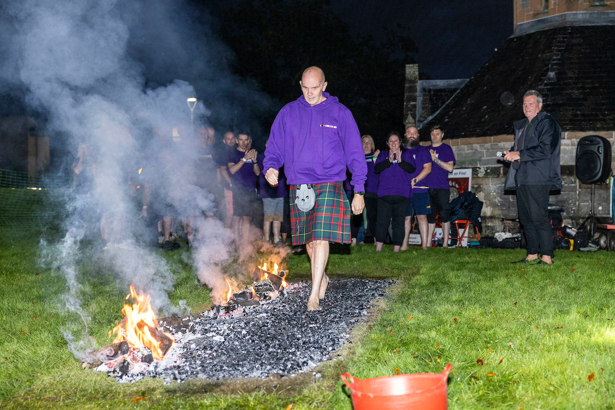 A person walks barefoot on burning coals, feeling the heat with every step, while a group of people watches under the night sky.