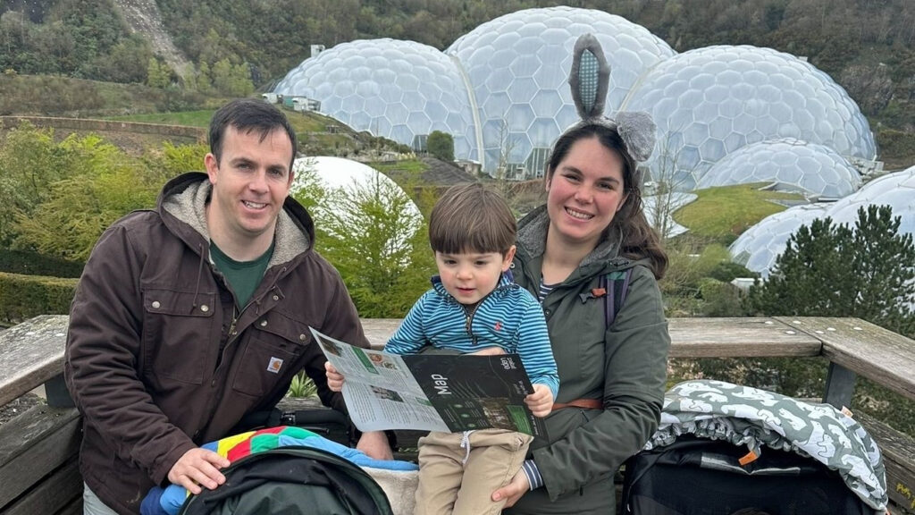 A military family of three poses outdoors, with a child holding a map. Behind them, geodesic domes stand as beacons of hope amidst the lush greenery.