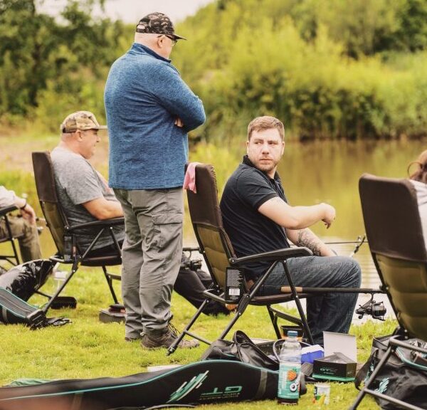 Five people are seated on folding chairs by a lakeside, engaging in fishing.