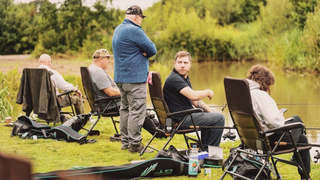 Five people are seated on folding chairs by a lakeside, engaging in fishing.