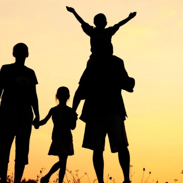 Silhouette of a family of four at sunset, with one child on an adult's shoulders and another child holding hands, evoking a scene reminiscent of Greenwich Hospital's community spirit.