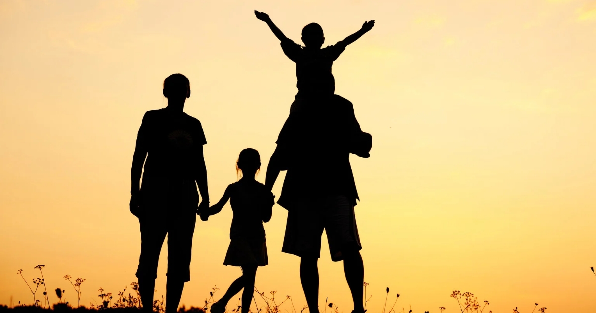 Silhouette of a family of four at sunset, with one child on an adult's shoulders and another child holding hands, evoking a scene reminiscent of Greenwich Hospital's community spirit.