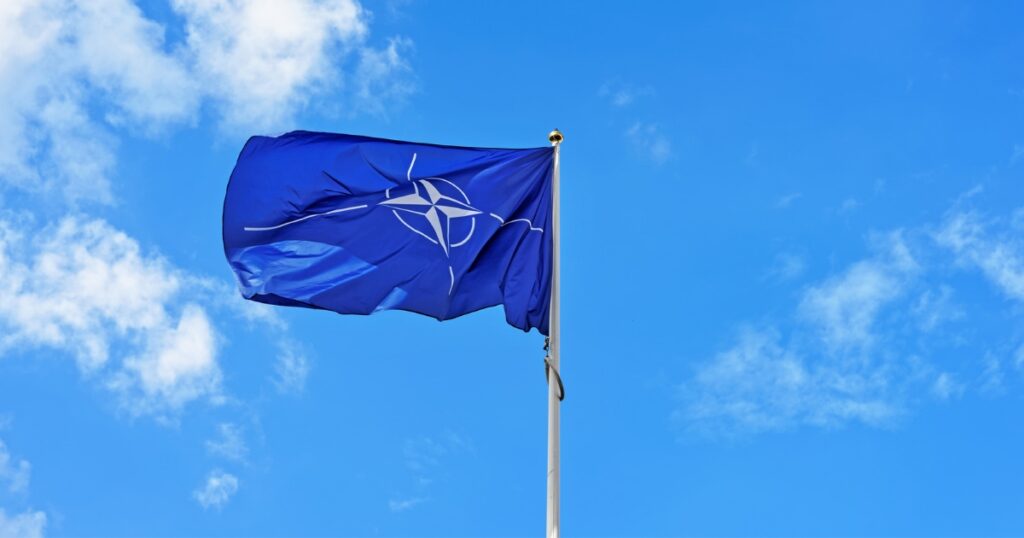 The NATO flag waves proudly on a flagpole against a clear blue sky, as UK troops participate in a significant NATO exercise in Eastern Europe.