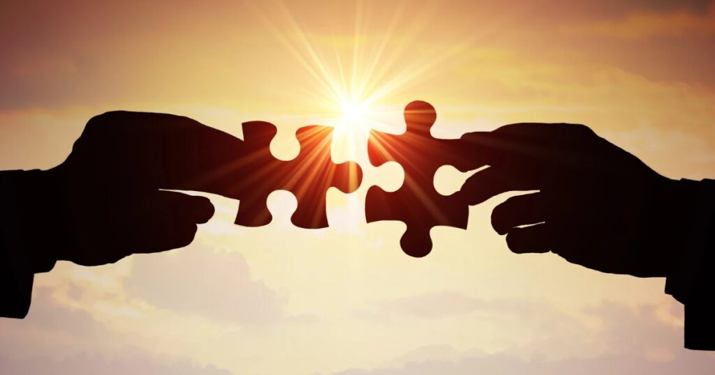 Two silhouetted hands holding puzzle pieces against a sunset background, with the sun shining between the pieces.