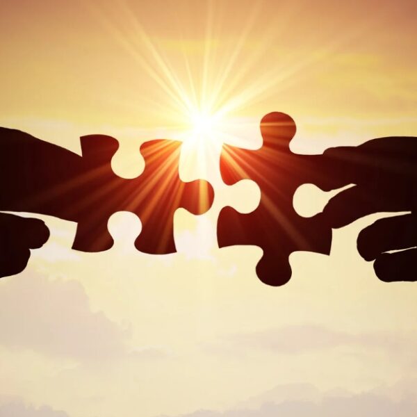 Two silhouetted hands holding puzzle pieces against a sunset background, with the sun shining between the pieces.