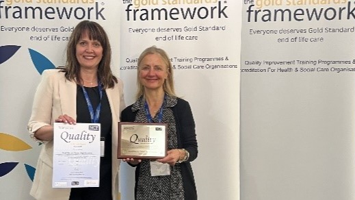 Two women standing, holding awards in front of banners for the Gold Standards Framework, related to end-of-life care quality.