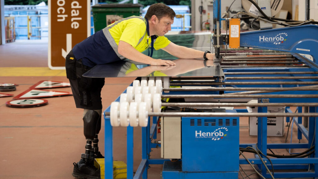 In a bustling workshop, a man with a prosthetic leg skillfully operates machinery, shaping metal under the banner of Scotland’s Bravest Manufacturing Company. Clad in a high-visibility shirt, his work contributes to ongoing research into impactful craftsmanship and rehabilitation.