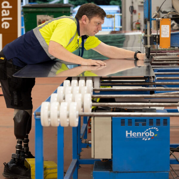 In a bustling workshop, a man with a prosthetic leg skillfully operates machinery, shaping metal under the banner of Scotland’s Bravest Manufacturing Company. Clad in a high-visibility shirt, his work contributes to ongoing research into impactful craftsmanship and rehabilitation.