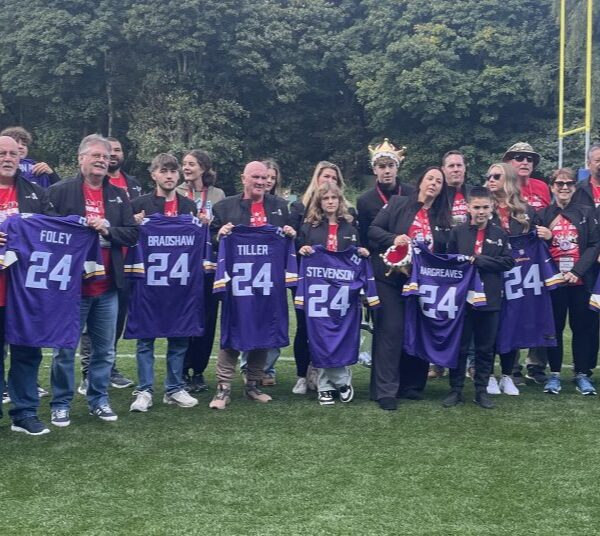 A group of people stands on a field holding purple jerseys with the number 24 on them, posing for a photo. Trees are visible in the background.