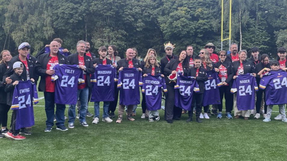 A group of people stands on a field holding purple jerseys with the number 24 on them, posing for a photo. Trees are visible in the background.