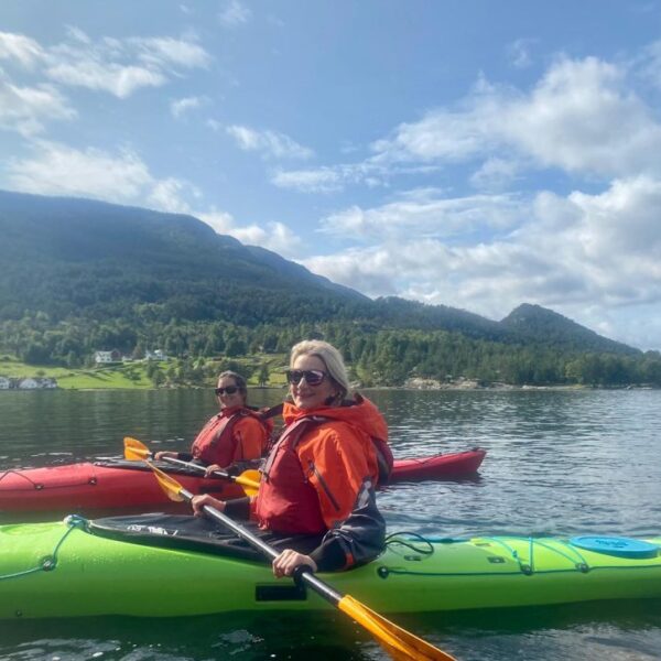 Two people in orange life jackets kayaking on calm water with mountains and trees in the background under a partly cloudy sky.