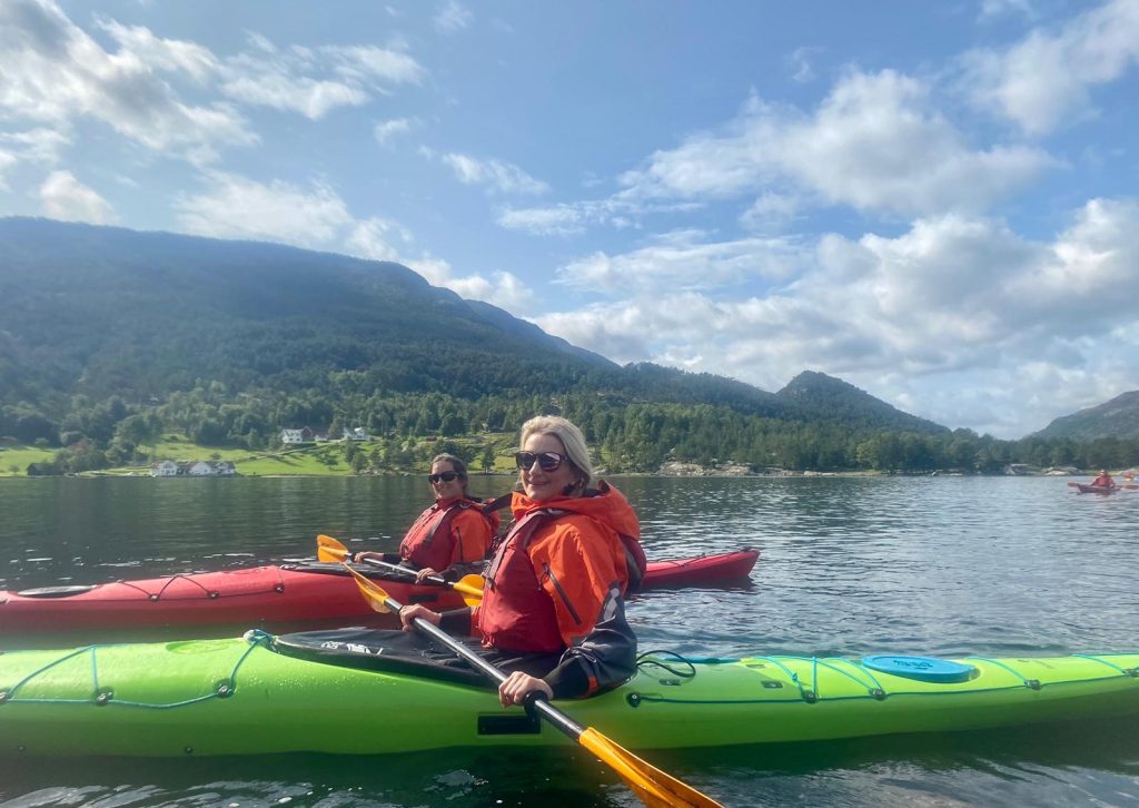 Two people in orange life jackets kayaking on calm water with mountains and trees in the background under a partly cloudy sky.