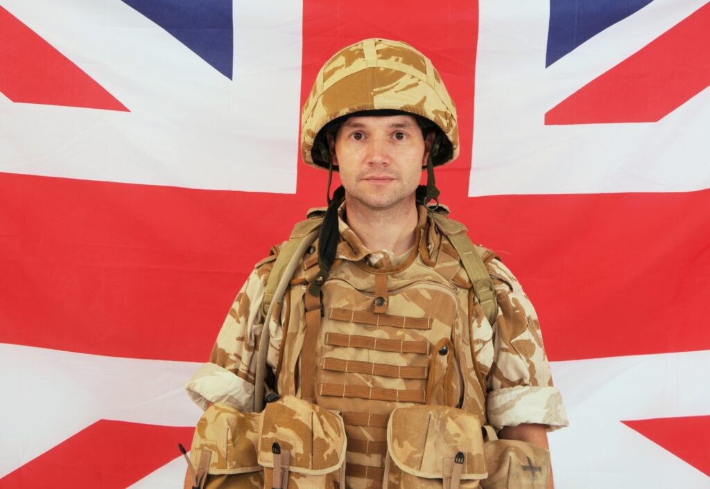 A soldier in camouflage uniform and helmet stands in front of a Union Jack flag.