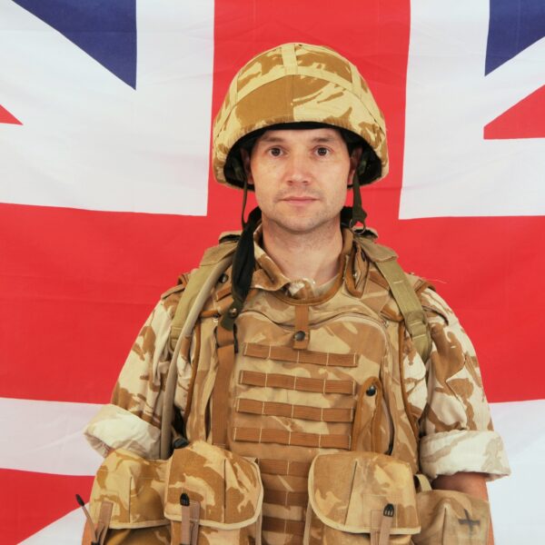 A soldier in camouflage uniform and helmet stands in front of a Union Jack flag.