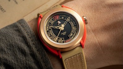 Close-up of a wristwatch with a historically unique dial featuring various text labels. The watch has a tan strap and a red bezel, worn on the wrist of someone supporting the Veterans Foundation.