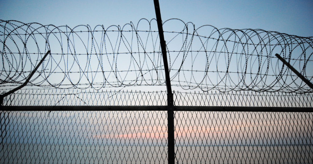 Barbed wire fencing against a twilight sky, with coiled razor wire along the top.