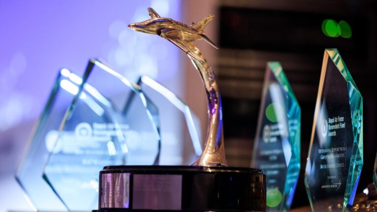 A collection of glass awards is displayed on a table, with one prominent award shaped like an aircraft on a stand in the foreground, celebrating the RAF Benevolent Fund. This remarkable display sets the stage for Awards 2025, capturing elegance and honor in each piece.
