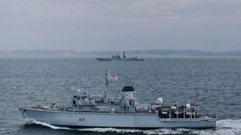 Two naval ships navigate the ocean; a foreground vessel displays a prominent red and white flag, while a larger ship from the Royal Navy looms in the distance. The sky is overcast, and land is faintly visible on the horizon.