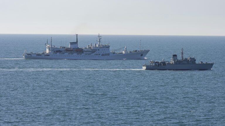 Two ships on the ocean: a large white vessel sails parallel with a smaller gray military ship in the foreground, possibly part of the Royal Navy patrolling UK waters.