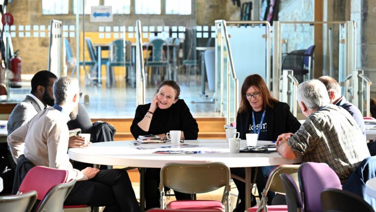 People sitting around a table in a well-lit room, engaged in empowering discussions. Various papers and cups are on the table.